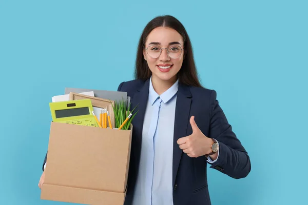 Happy unemployed woman with box of personal office belongings showing thumbs up on light blue background