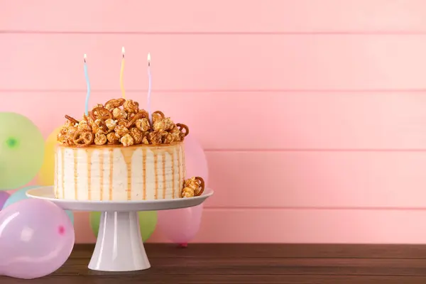 Caramel drip cake decorated with popcorn and pretzels near balloons on wooden table, space for text