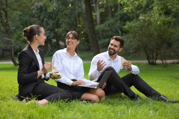 Bilgisayarlı mutlu meslektaşlar parkta yeşil çimlerde iş yemeği yiyorlar.