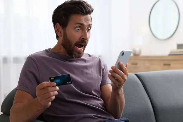 Shocked man with credit card using smartphone on sofa at home. Be careful - fraud