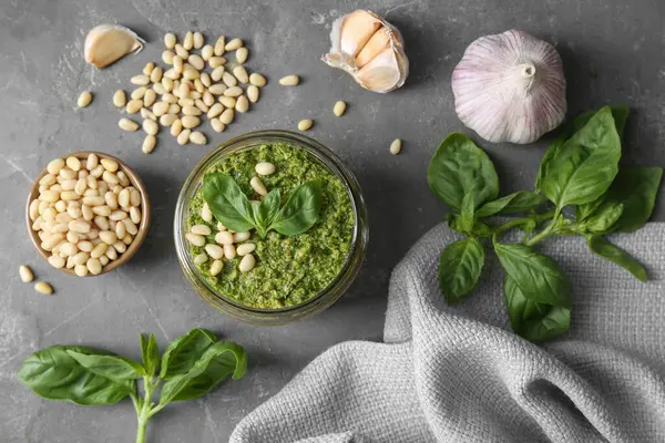 Jar of delicious pesto sauce and ingredients on grey table, flat lay