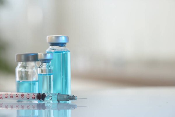 Glass vials with light blue medication and syringe on white table, closeup. Space for text