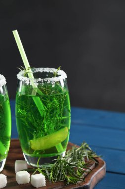 Bir bardak ev yapımı taze tarhun içeceği, dallar ve şeker küpleri mavi ahşap masada.