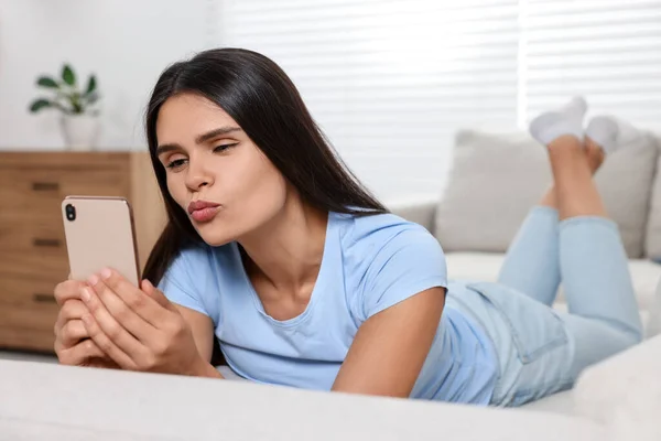Young woman having video chat via smartphone and sending air kiss on sofa in living room
