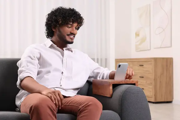 Happy man using smartphone on sofa armrest wooden table at home