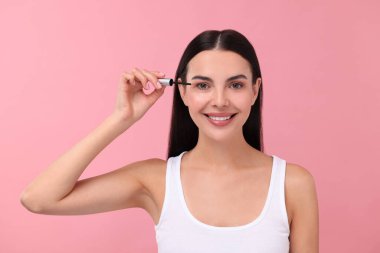 Güzel bir kadın pembe arka planda kirpiklerine serum sürüyor. Kozmetik ürünü