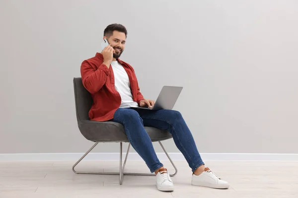 Handsome man with laptop talking on smartphone while sitting in armchair near light grey wall indoors