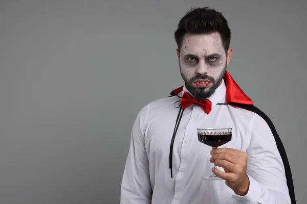 Man in scary vampire costume with fangs and glass of wine on light grey background, space for text. Halloween celebration
