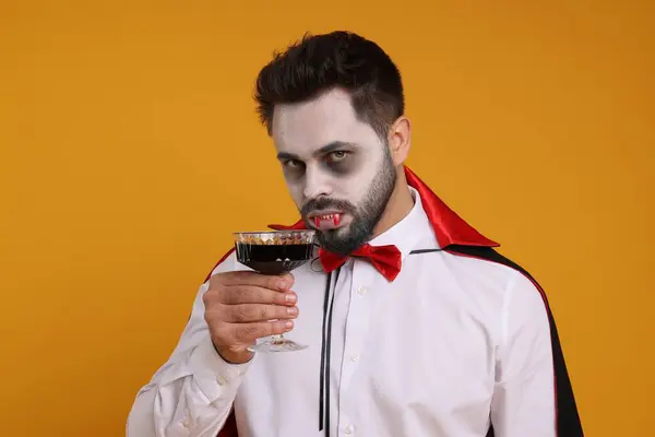 Man in scary vampire costume with fangs and glass of wine on orange background. Halloween celebration