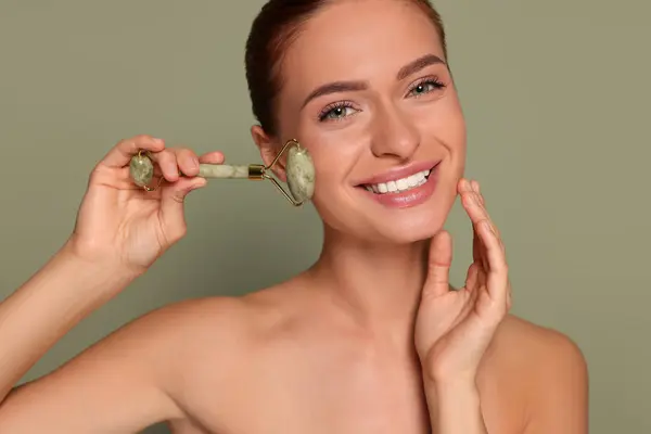 stock image Young woman massaging her face with jade roller on green background