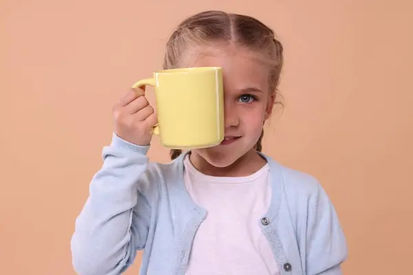 Happy girl covering eye with yellow ceramic mug on beige background
