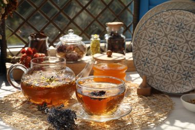 Aromatik çay, farklı kuru bitkiler ve pencerenin yanındaki beyaz ahşap masada bal.