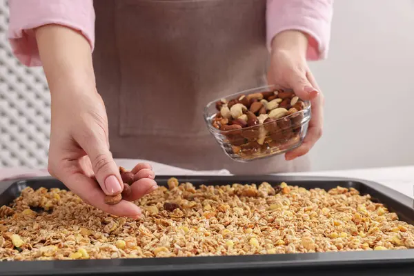 Making granola. Woman adding nuts onto baking tray with mixture of oat flakes and other ingredients at table in kitchen, closeup
