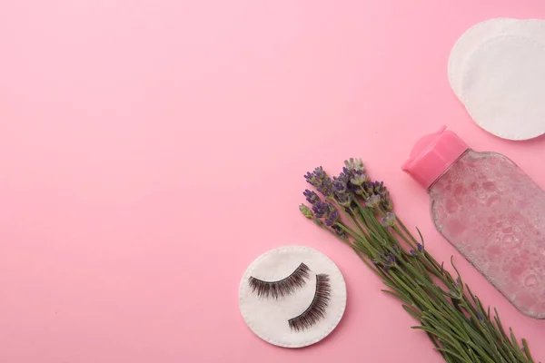 Bottle of makeup remover, lavender, cotton pads and false eyelashes on pink background, flat lay. Space for text