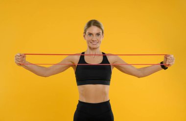 Turuncu arka planda elastik direnç bandıyla egzersiz yapan kadın.