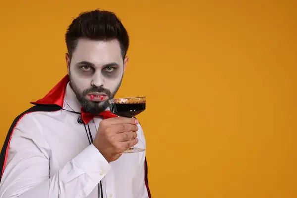 Man in scary vampire costume with fangs and glass of wine on orange background, space for text. Halloween celebration