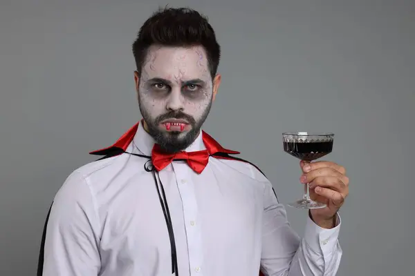 Man in scary vampire costume with fangs and glass of wine on light grey background. Halloween celebration