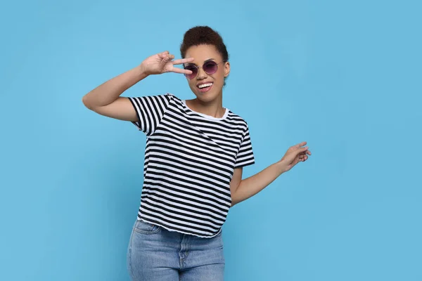 Happy young woman in stylish sunglasses dancing on light blue background