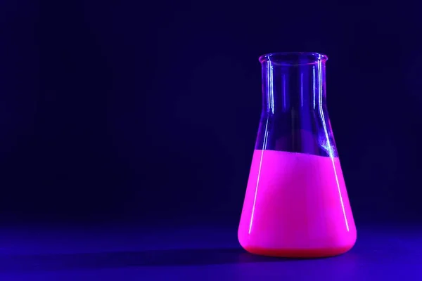 Laboratory flask with luminous liquid on dark blue background, space for text