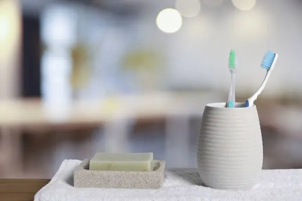 Plastic toothbrushes in holder, soap and towel on wooden table against blurred background, space for text
