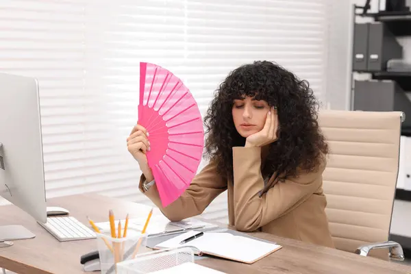 stock image Young businesswoman waving hand fan to cool herself at table in office