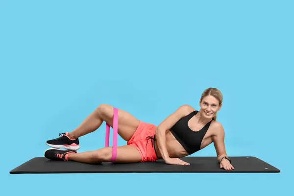 Woman exercising with elastic resistance band on fitness mat against light blue background