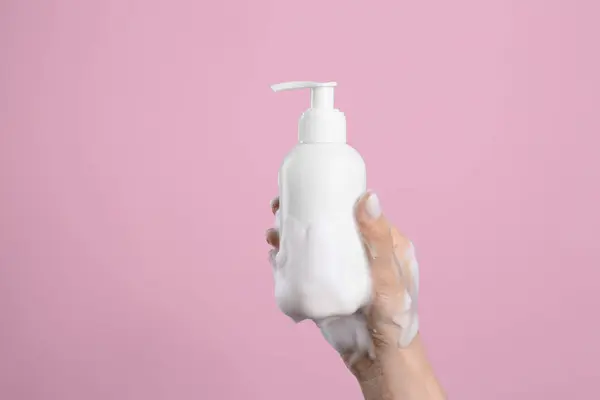 Woman with foam on hand holding bottle of skin cleanser against pink background, closeup. Cosmetic product
