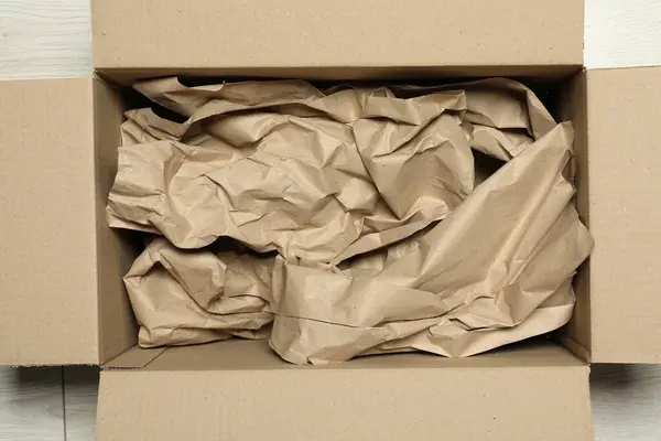 stock image One open cardboard box with crumpled paper on white wooden floor, top view