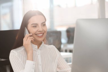 Ofiste akıllı telefonla konuşurken modern bilgisayar kullanan mutlu bir kadın.