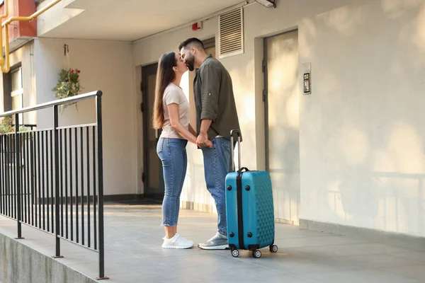 Long-distance relationship. Suitcase and beautiful young couple kissing near house entrance outdoors