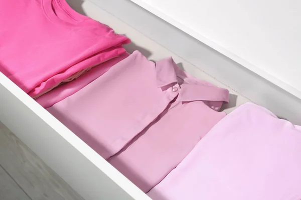 Many folded pink clothes in white chest of drawers indoors, closeup