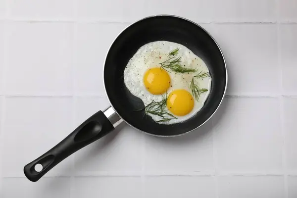 Frying pan with tasty cooked eggs and dill on white tiled table, top view