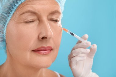 Doktor, açık mavi arka planda yaşlı kadına yüz enjeksiyonu yapıyor. Yakın plan. Estetik ameliyat