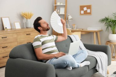 Sakallı ve elinde beyaz el yelpazesi olan adam evdeki kanepede serinliyor.