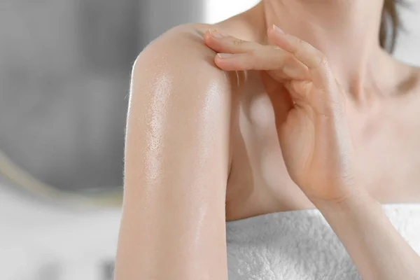 Woman applying body oil onto shoulder in bathroom, closeup