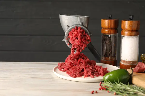 Metal meat grinder with minced beef, products and spices on light wooden table. Space for text