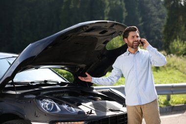 Stresli bir adam dışarıda bozuk bir arabanın yanında akıllı telefondan konuşuyor.