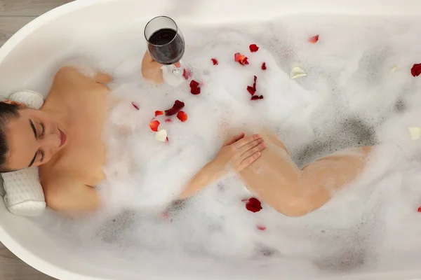 Woman with glass of wine taking bath in tub with foam and rose petals, top view
