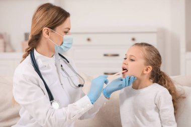 Tıbbi maskeli doktor, kızın ağız boşluğunu içeride dil bastırıcı ile inceliyor.