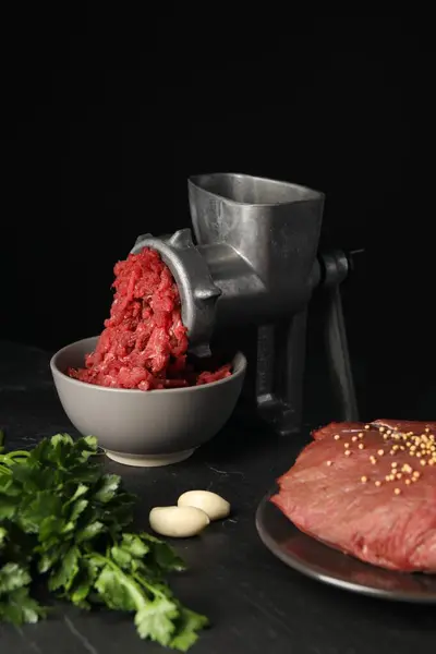 Metal meat grinder with beef mince and spices on dark textured table against black background
