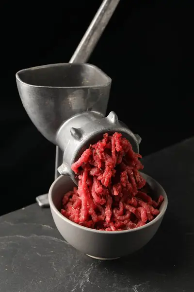 Metal meat grinder with beef mince on dark textured table against black background