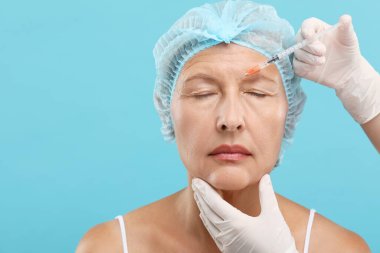 Doktor açık mavi arka planda yaşlı kadına yüz enjeksiyonu yapıyor. Estetik ameliyat