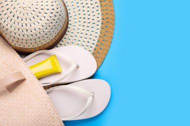 Hasır şapka, çanta, güneş kremi ve açık mavi arka planda parmak arası terlikler, düz yatış ve metin için boşluk. Plaj aksesuarları