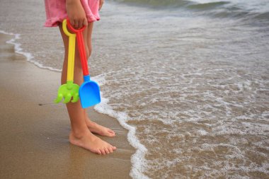 Plastik oyuncakları olan küçük bir kız sahile yakın kumsalda duruyor. Metin için boşluk