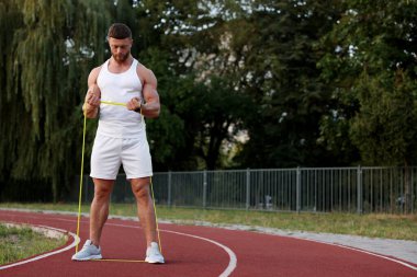 Stadyumda elastik direniş bandıyla egzersiz yapan kaslı adam.