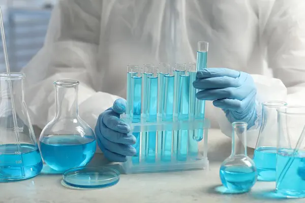 Scientist taking test tube with light blue liquid at white table in laboratory, closeup
