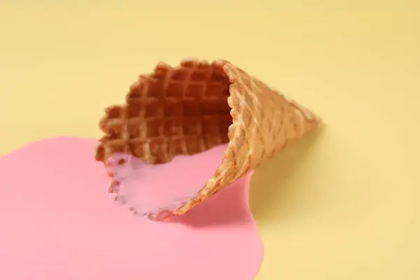 Melted ice cream and wafer cone on pale yellow background, closeup