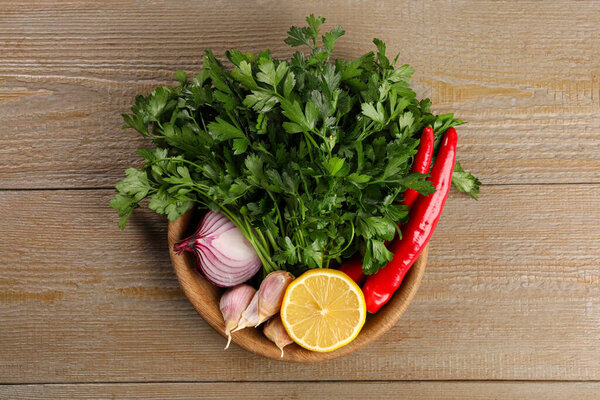 Bowl with fresh green parsley, chili peppers, lemon, onion and garlic on wooden table, top view