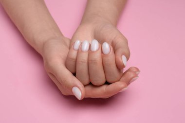 Manikürlü ellerini pembe arka planda beyaz tırnak cilası ile gösteren bir kadın.