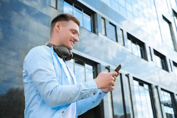 Smiling man with headphones using smartphone near building outdoors. Space for text